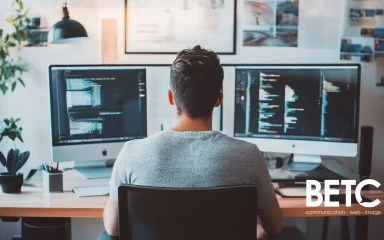 Développeur travaillant sur deux écrans avec du code à l'écran, logo de l'agence BETC en bas à droite.
