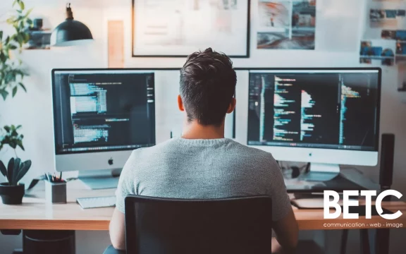 Développeur travaillant sur deux écrans avec du code à l'écran, logo de l'agence BETC en bas à droite.