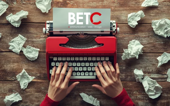 Mains tapant sur une machine à écrire rouge avec le logo BETC communication sur une feuille, entourée de papiers froissés sur une table en bois.